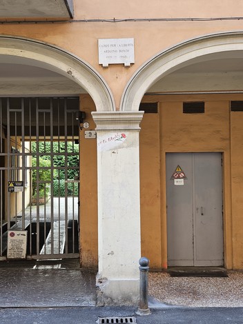 Lapide commemorativa su un muro in una zona urbana, con archi in muratura e ingresso a un cortile.