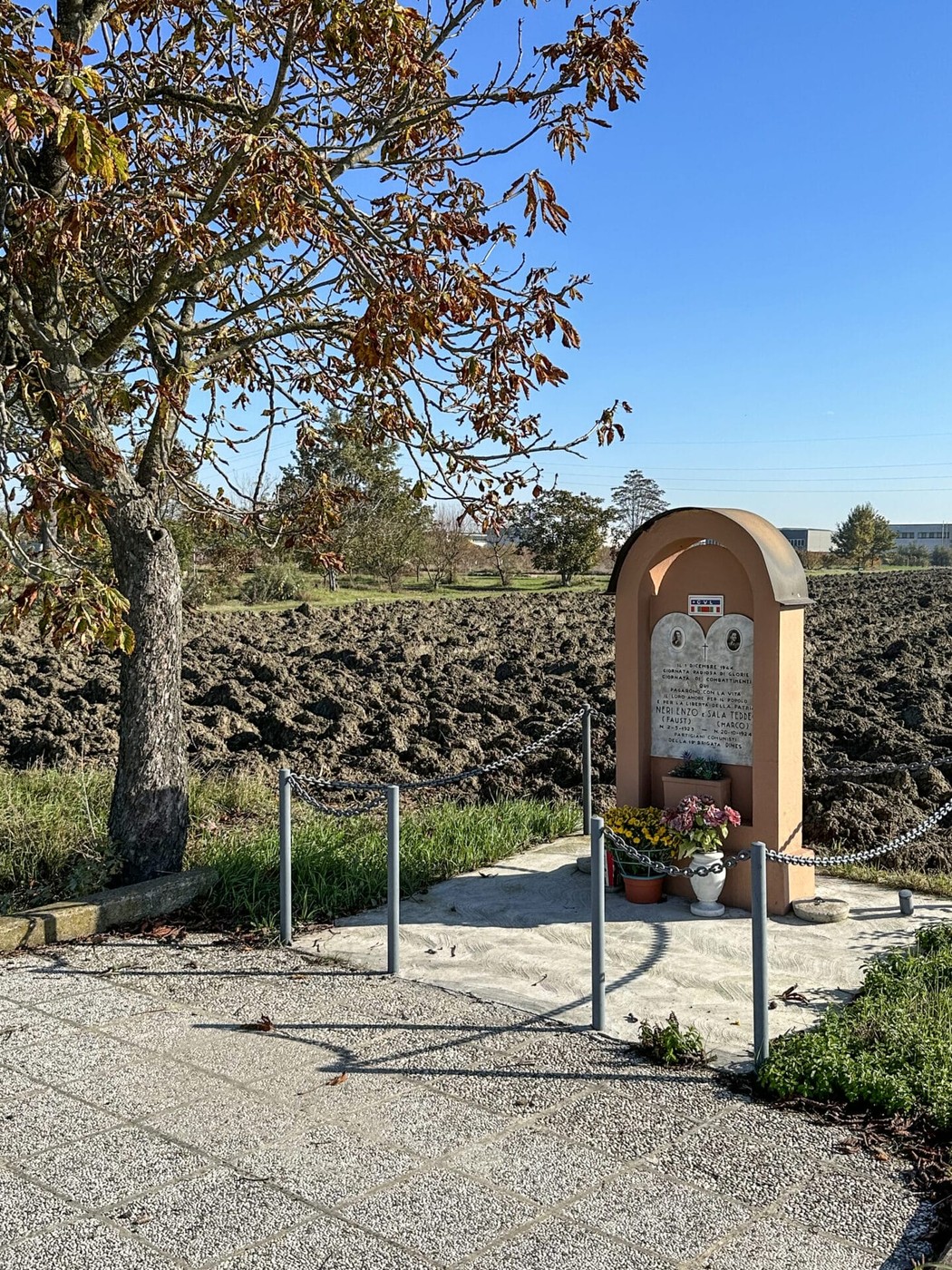 Monumento a Neri e Sala, situato in un paesaggio rurale, con piante e una recinzione semplice.
