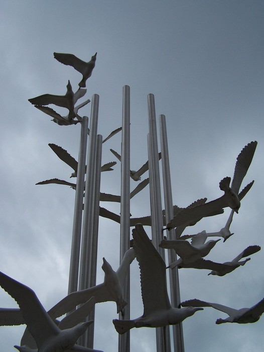 Monumento del sessantesimo, con uccelli argentati in volo e pilastri metallici, simbolo di libertà e progresso.