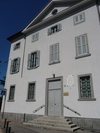 Lapide commemorativa su un edificio con architettura bianca e dettagli grigi, in contesto urbano.