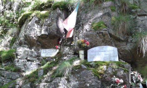 Lapidi commemorative all'Alpe Morcei, con fiori e bandiera tricolore, in un ambiente naturale suggestivo.