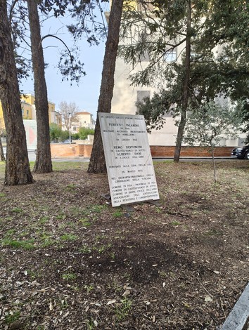 Stele commemorativa tra alberi, con edifici sullo sfondo, in un'area verde.