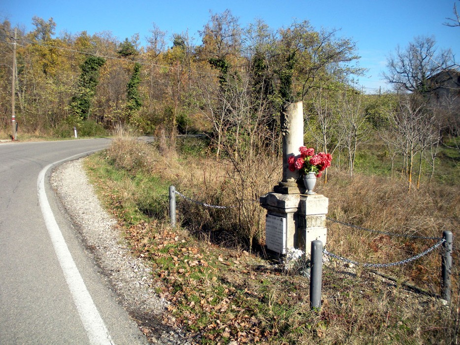 Cippo commemorativo con fiori, situato lungo una strada in un paesaggio naturale.