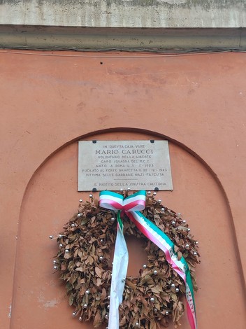 Lapide commemorativa di Mario Carucci, decorata con una corona di foglie e nastri, su parete di colore arancio.