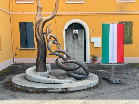 Scultura dell'Albero della Memoria, con bandiera italiana e targa commemorativa su sfondo giallo.