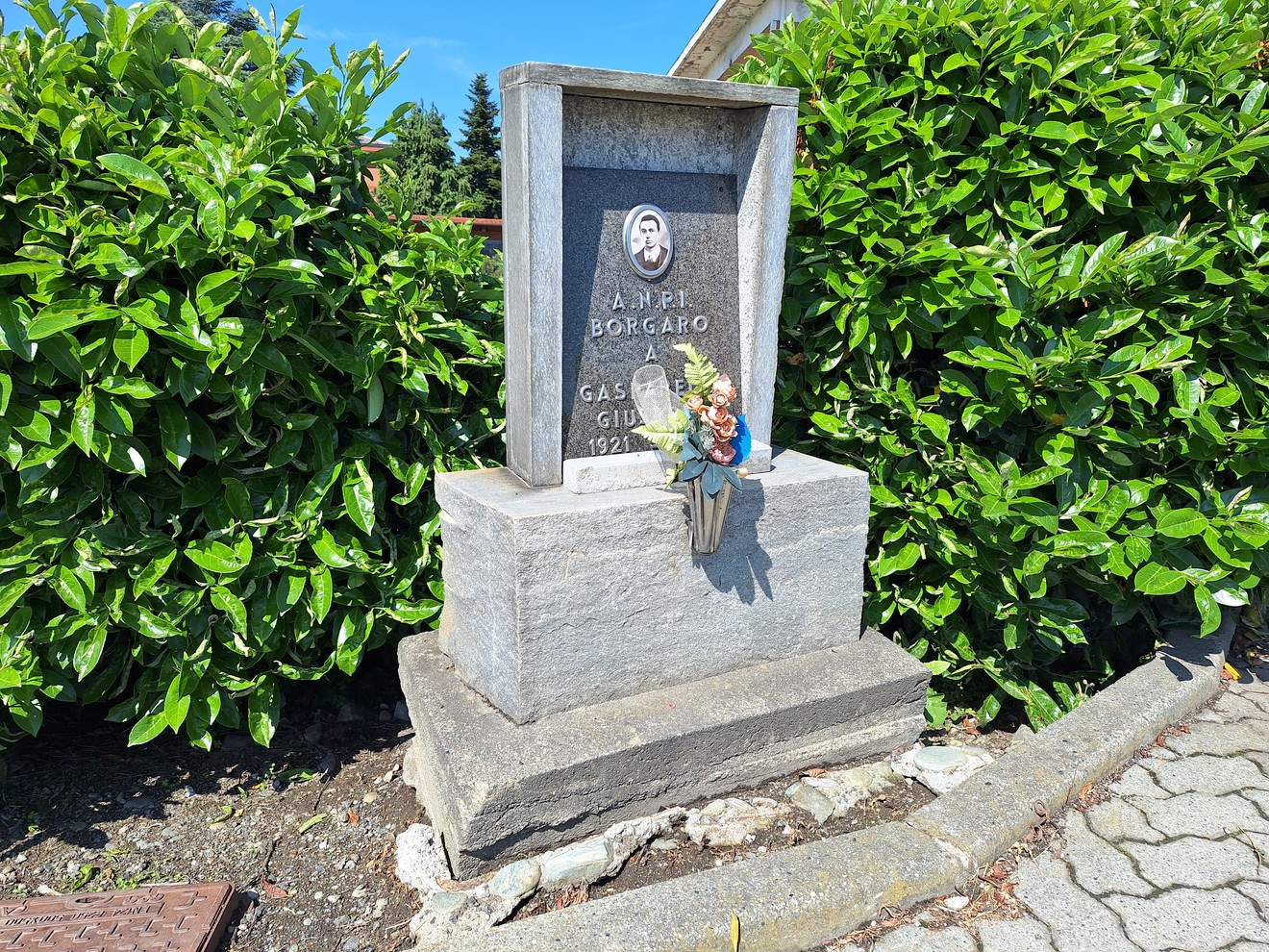 Cippo in pietra dedicato a Gasparello, con figura decorativa e fiori, immerso nel verde circostante.