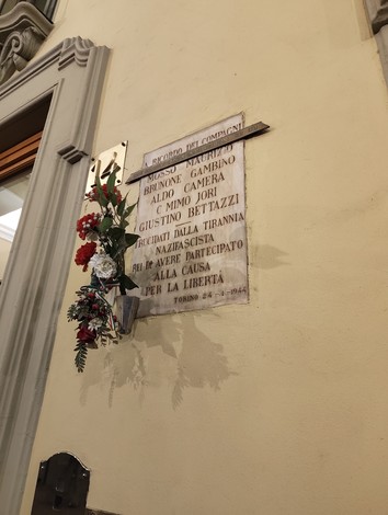 Lapide commemorativa con nomi di caduti, adornata da fiori, su muro in ambiente urbano.