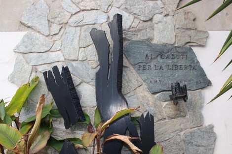 Monumento ai caduti per la Libertà, con elementi in pietra e legno scuro, circondato da piante verdi.