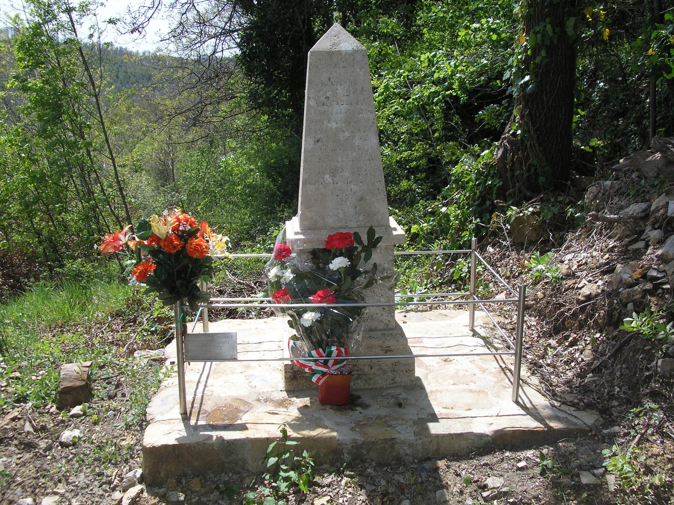 Cippo commemorativo con fiori, dedicato a Ghiribelli, Malossi e Tompetrini, immerso nella natura.
