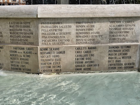 Fontana Monumentale con incisi nomi su una superficie rocciosa, spruzzi d'acqua e riflessi sul pelo dell'acqua.