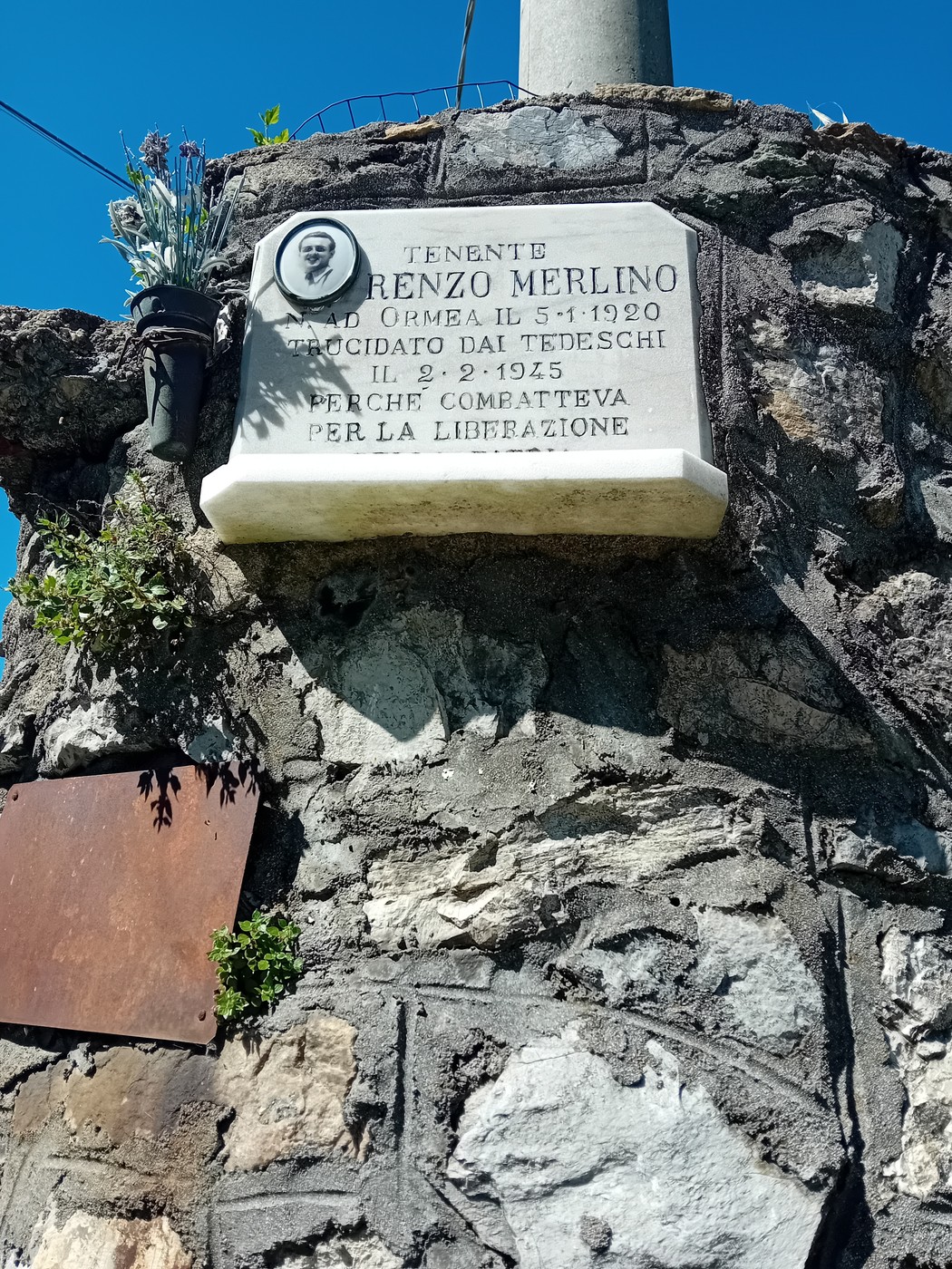 Lapide commemorativa dedicata a Lorenzo Merlino, situata su una roccia, con fiori e sotto un cielo azzurro.