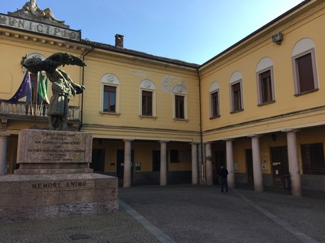 Lapide ai Caduti in un contesto urbano, con un monumento e l'edificio municipale sullo sfondo.