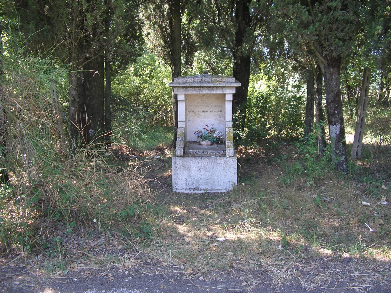 Tabernacolo a Rotelli immerso nella vegetazione, con un'edicola che custodisce fiori e segni di devozione.