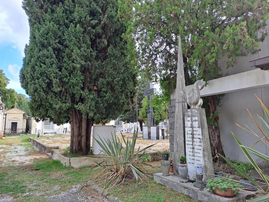 Cippo in pietra con iscrizione e statua, tra alberi e vegetazione in un'area cimiteriale.