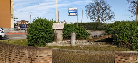 Stele commemorativa ai Caduti di Le Budrie, circondata da vegetazione, con segnaletica stradale sullo sfondo.