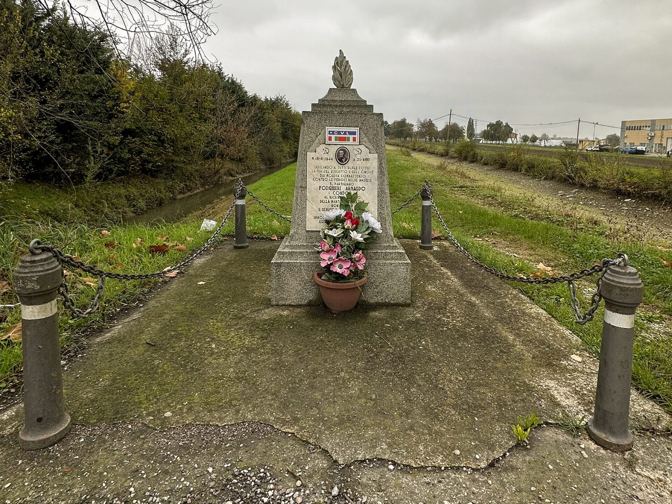 Cippo commemorativo a Forghieri, circondato da fiori e catene, in un contesto verde e rurale.
