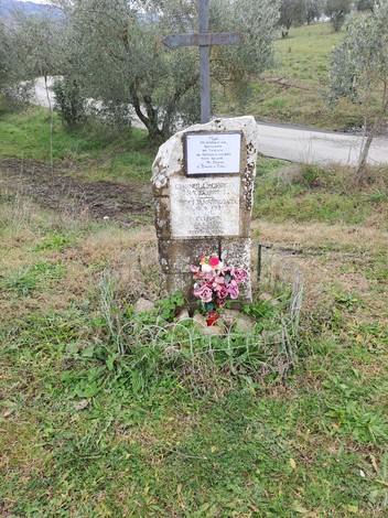 Cippo commemorativo con fiori, situato in un paesaggio rurale e circondato da ulivi.