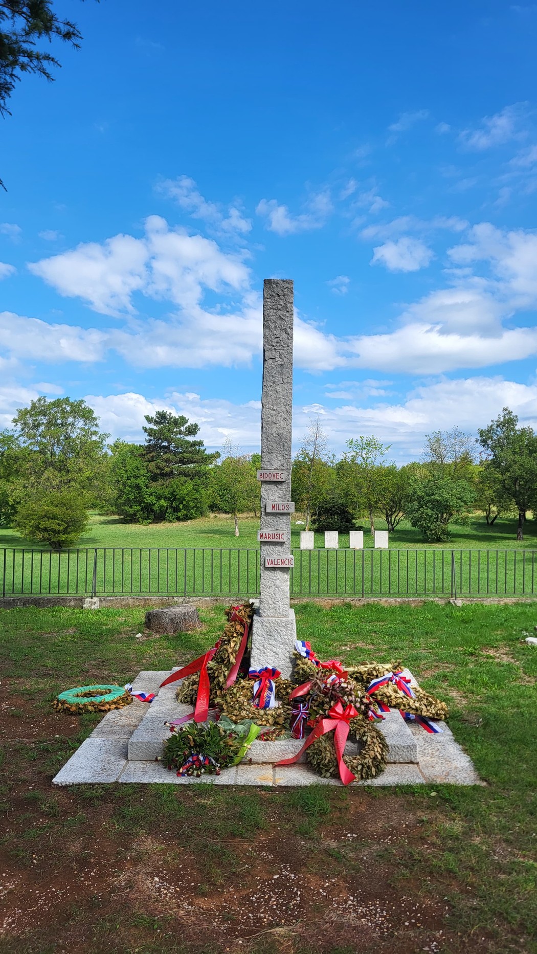 Monumento in pietra a forma di stele, con targhe incise, circondato da corone di fiori nel prato.
