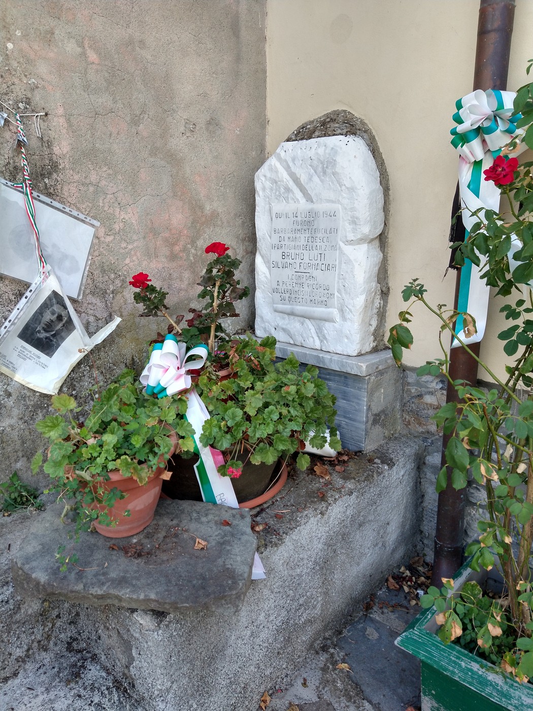 Cippo commemorativo a Luti e Fornaciari, adornato con fiori e nastri tricolore, in un angolo di memoria.