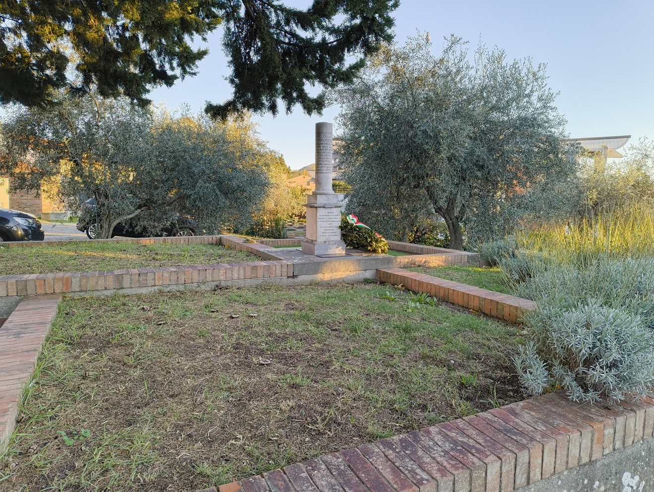 Cippo commemorativo dedicato ai caduti della Seconda Guerra Mondiale, circondato da olivi e vegetazione.