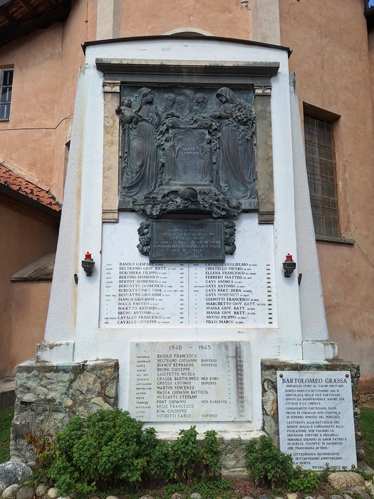 Monumento in bronzo e marmo con iscrizioni, fissato sulla parete di un edificio e circondato da vegetazione.