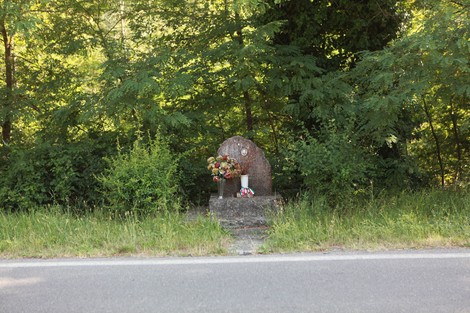 Monumento a Masini, circondato da vegetazione, con fiori e lumi commemorativi ai piedi.