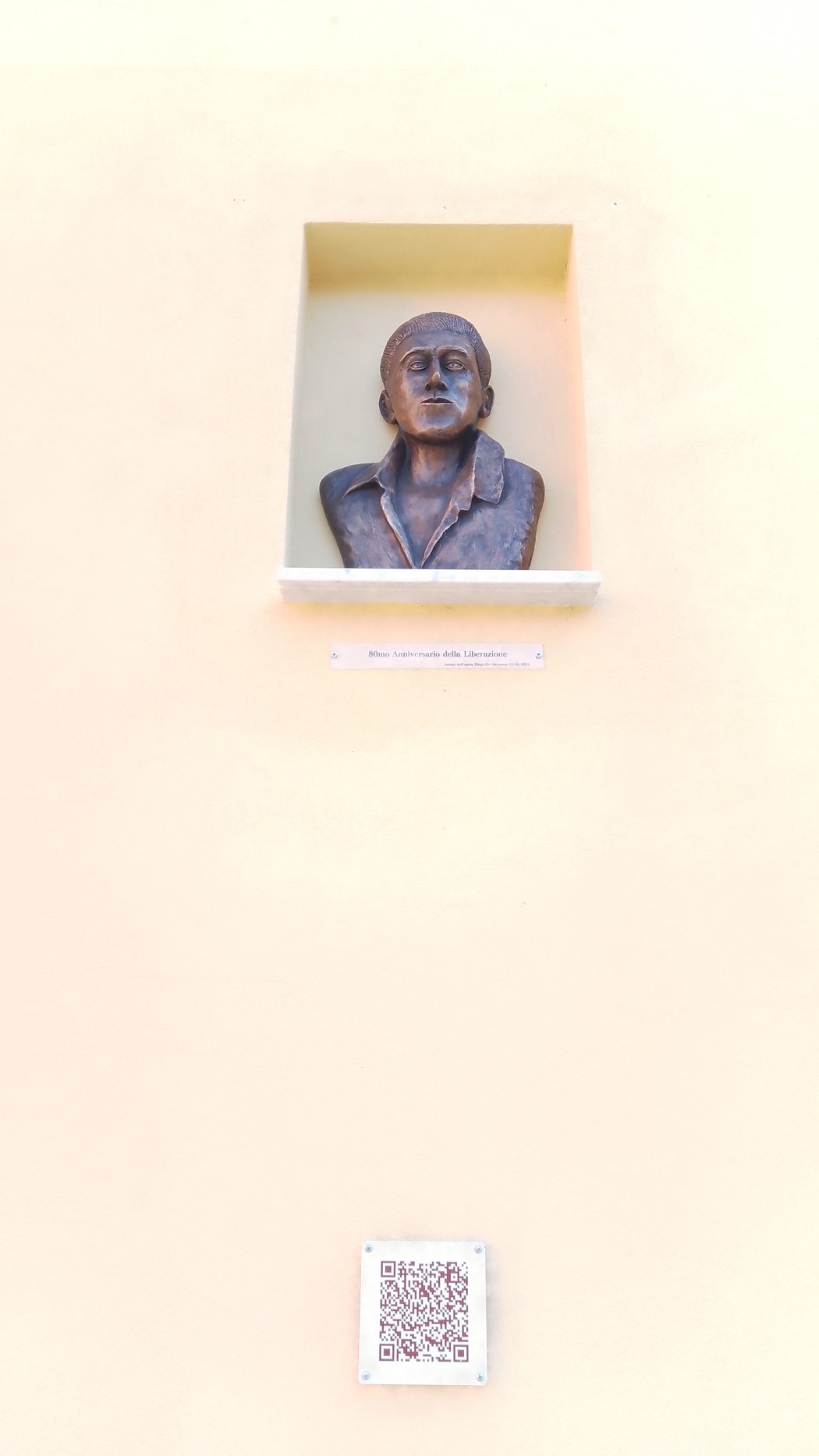 Busto in bronzo su parete gialla, rappresentante un volto maschile, con QR code sottostante.