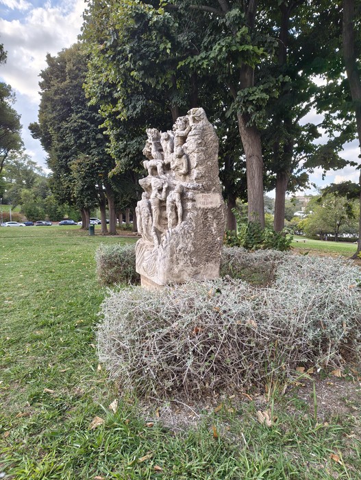 Monumento in pietra con figure scolpite e iscrizione, collocato in un’aiuola alberata.