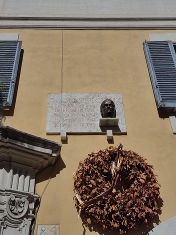 Lapide commemorativa con busto e ghirlanda, affissa su un muro giallo.
