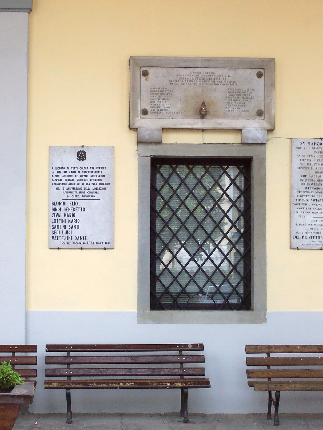 Lapide commemorativa in onore dei caduti, affissa su una parete con panchine sottostanti.