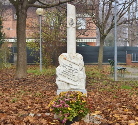 Cippo commemorativo a Goldoni, con iscrizioni e decorazioni floreali, immerso in un parco con foglie autunnali.
