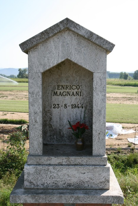 Tabernacolo dedicato a Enrico Magnani, con fiori e un paesaggio rurale sullo sfondo.