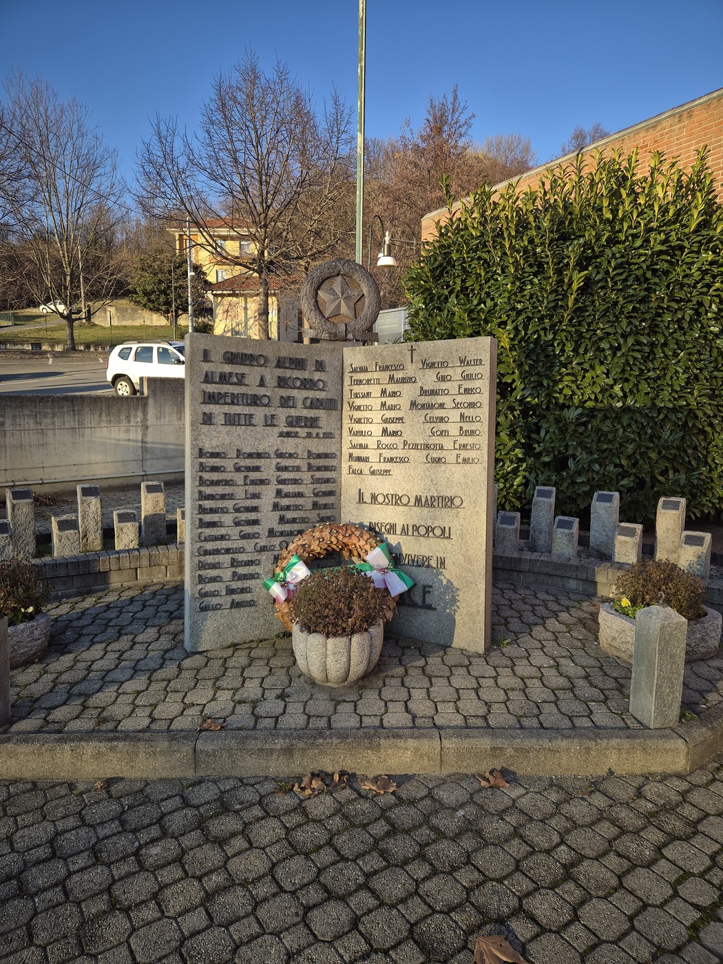 Monumento in pietra con lapidi incise e corona di fiori, in un'area pavimentata circondata da siepi.