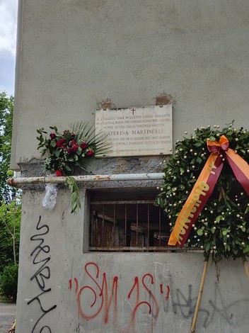 Lapide dedicata a Caterina Martinelli, adornata con fiori e una corona, in un contesto urbano.