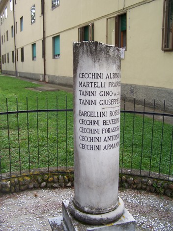 Cippo commemorativo con nomi incisi, situato in un'area verde circondata da una balaustra.