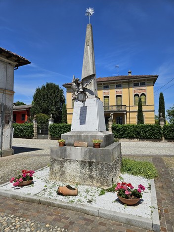 Monumento ai Caduti di Castelponzone, con obelisco e decorazioni, circondato da piante e fiori.