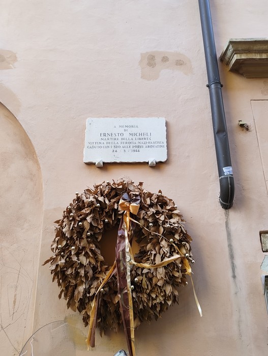 Lapide rettangolare in marmo bianco fissata a una parete, con iscrizione e corona commemorativa appesa sotto.