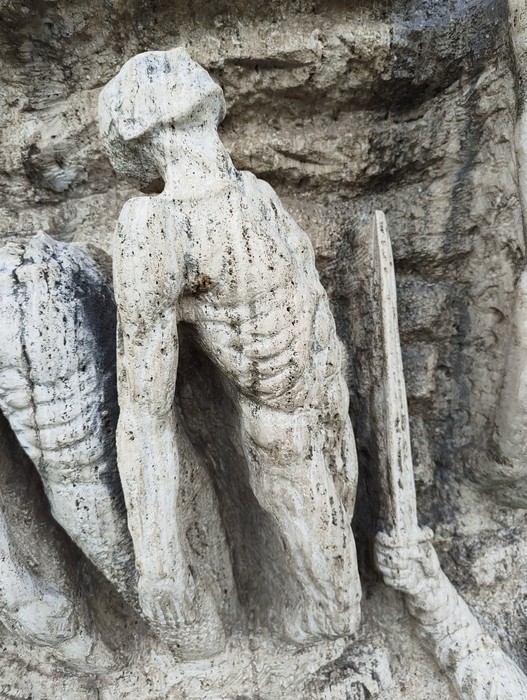 Scultura in pietra di una figura umana inginocchiata, inserita in una parete rocciosa e accanto a un fucile.
