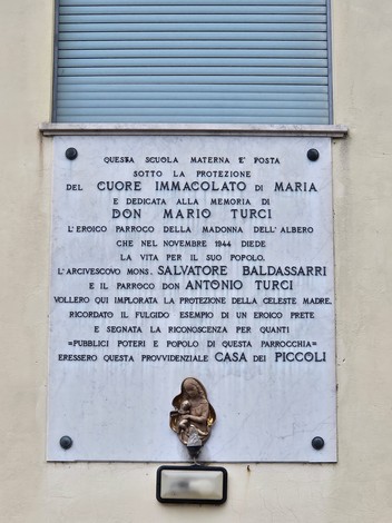 Lapide commemorativa dedicata a Don Mario Turci, con riferimento alla protezione di Maria e alla vita della comunità.