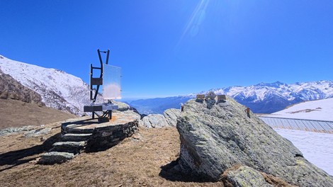 Struttura metallica con pannello su roccia in alta montagna, intorno vette innevate sotto cielo terso.