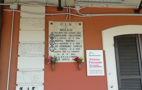 Lapide in marmo con iscrizione affissa su una parete esterna, accanto a vasi di fiori.