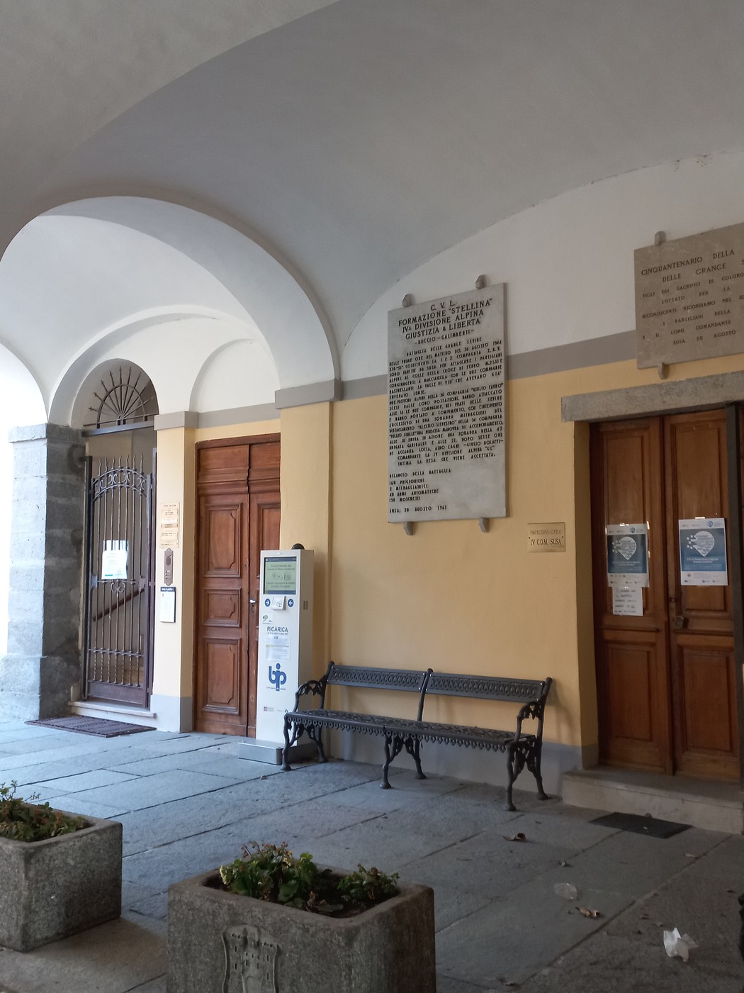 Lapide commemorativa della formazione "Stellina" in un contesto architettonico con porte e panchina.