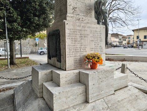 Monumento ai caduti con fiori, situato in un'area pubblica, circondato da alberi e strade.