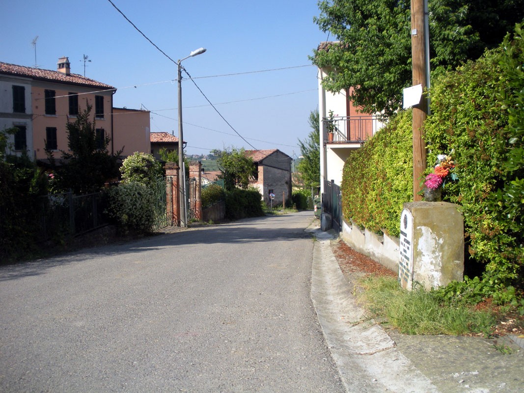 Cippo a Francescotti in un contesto rurale, con strada e case sullo sfondo verdeggiante.