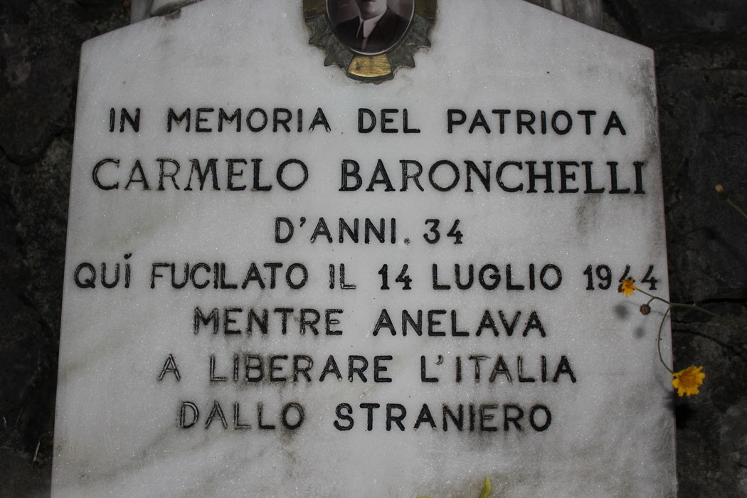 Lapide commemorativa dedicata a Carmelo Baronchelli, patriota fucilato nel 1944, immersa nella natura.