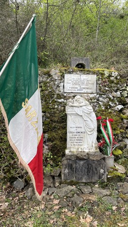 Monumento a Foschi con lapide e bandiera italiana, immerso nella natura.
