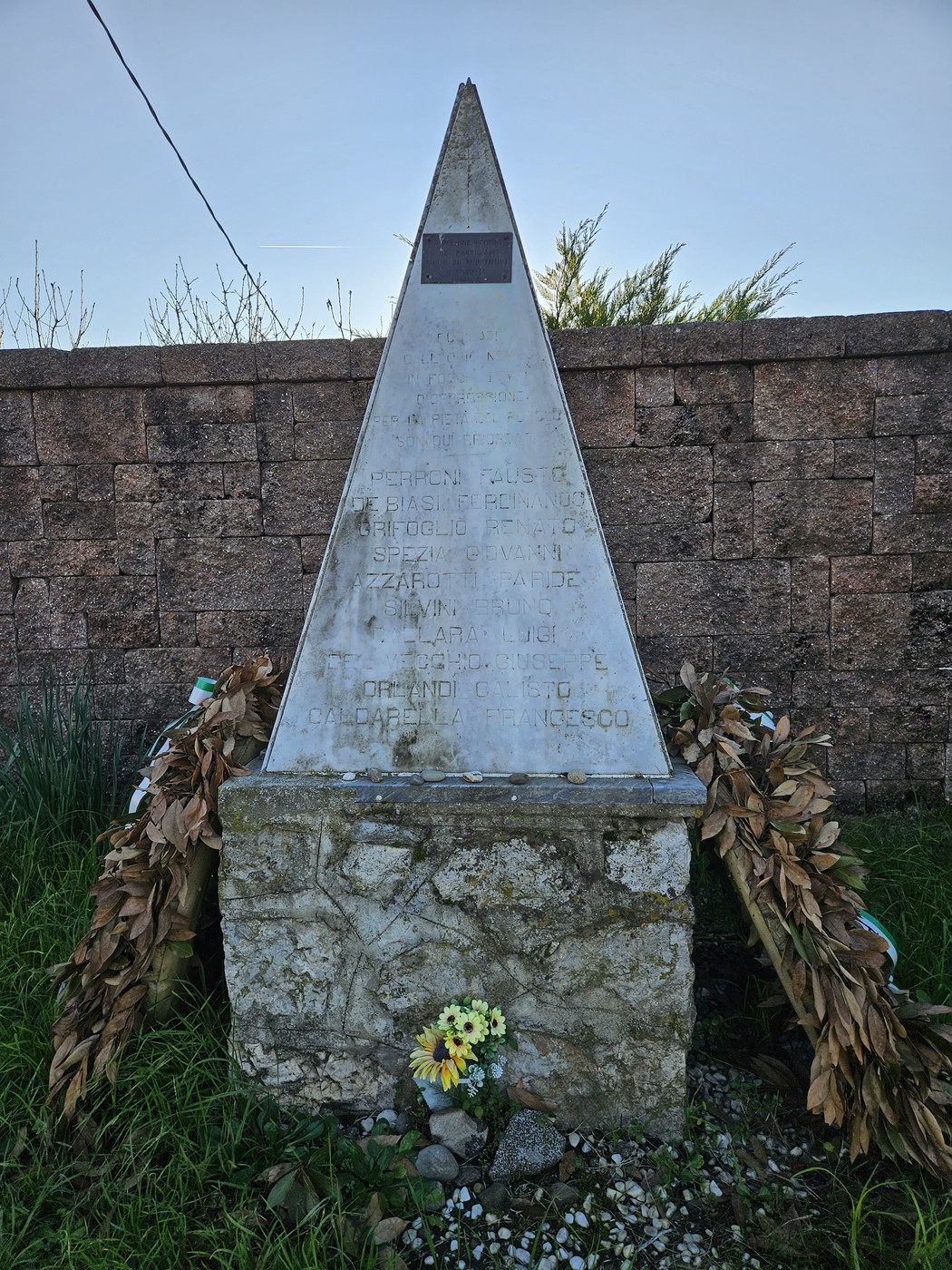 Monumento ai Martiri di Ressora, obelisco con decorazioni floreali e un muro di pietra sullo sfondo.