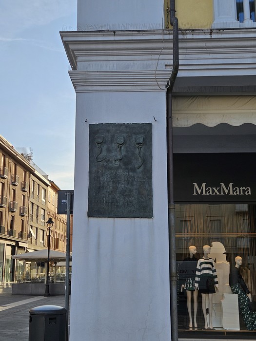 Targa commemorativa ai Tre Martiri, con rilievi su fondo scuro, affissa a un muro bianco in un'area urbana.
