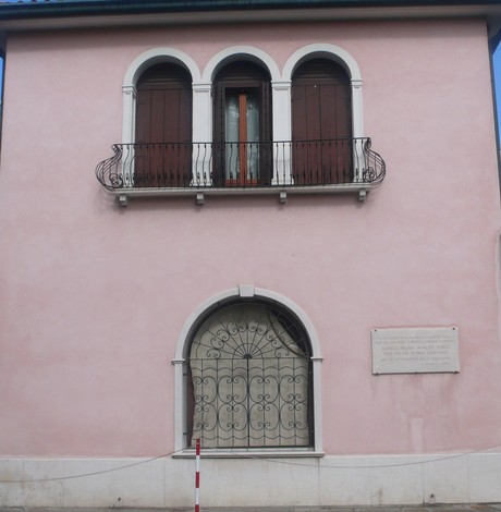 Lapide commemorativa su una facciata di un edificio rosa con balcone e decorazioni in ferro battuto.