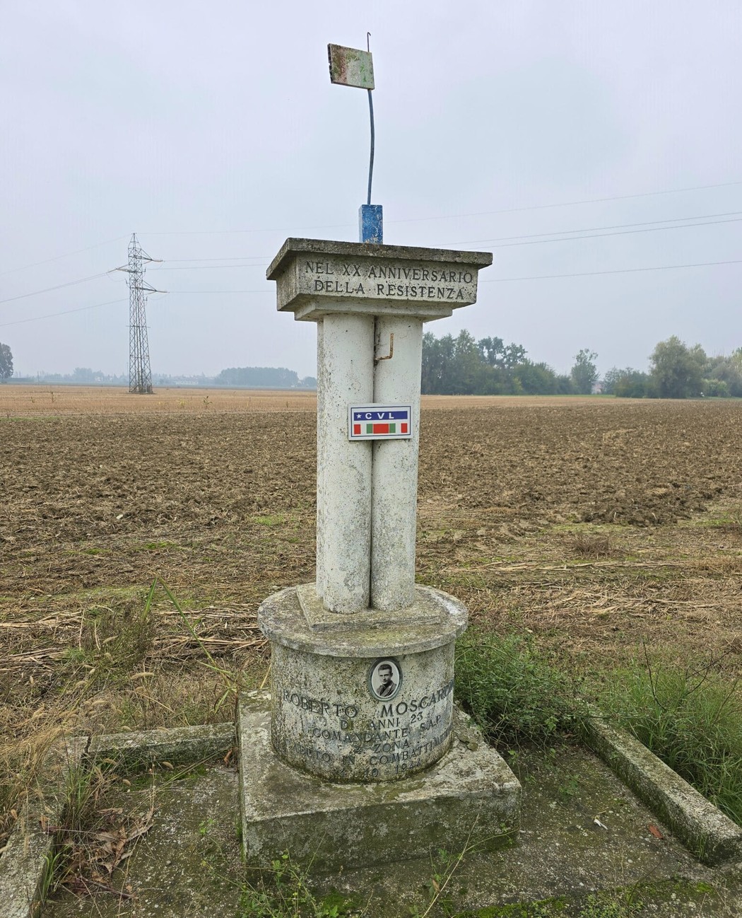 Cippo commemorativo a Moscardini, con bandiera e contesto rurale alle spalle.